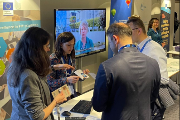 A photo of the PM² booth with two PM² team members showing PM² materials to two visitors