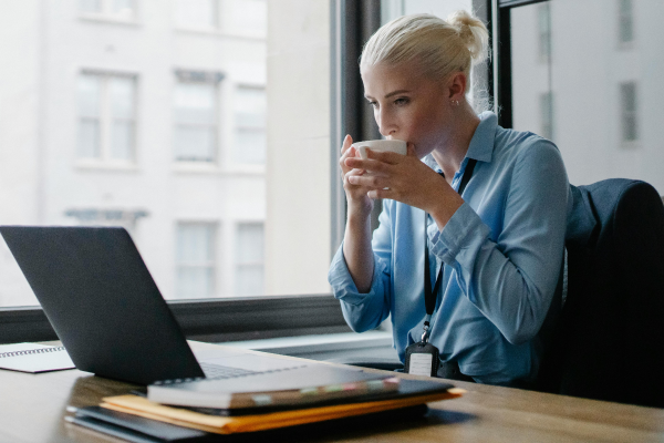 A professional woman sits at a desk with a laptop, sipping coffee while looking at the screen. 